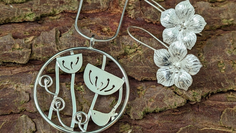 A silver pendant depicting a bird and seed heads and a pair of flower-shaped earrings rest against tree bark.