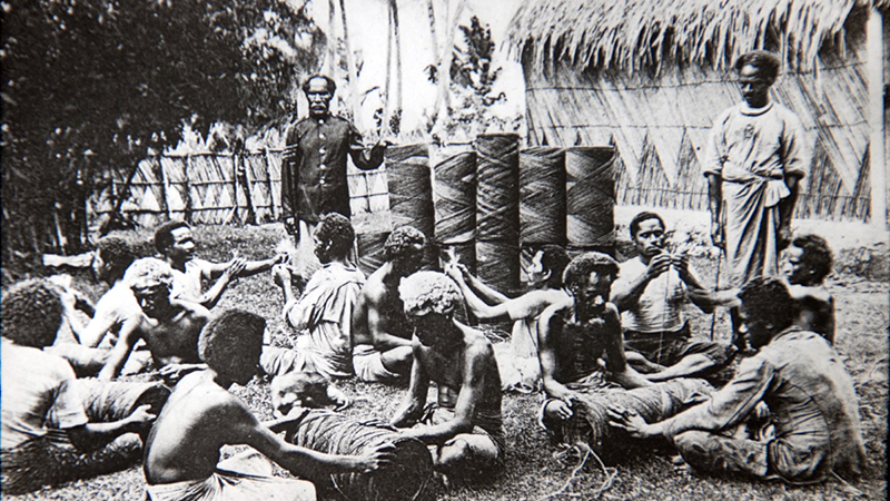 Men sit in front of coir coils, at work rolling more. Two men stand behind, overseeing the work.