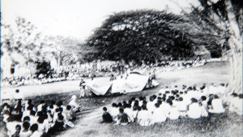 Slightly blurry black and white photograph of a ceremony, with many people sitting around a central performance