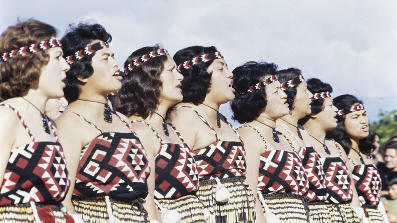 A group of 8 Māori women performing a dance. They are wearing tops, and headbands in black, red and white patterns, piupiu (skirts), and hei tiki (pendants). They are standing shoulder to shoulder with hands by their sides and appear to be vocalising.