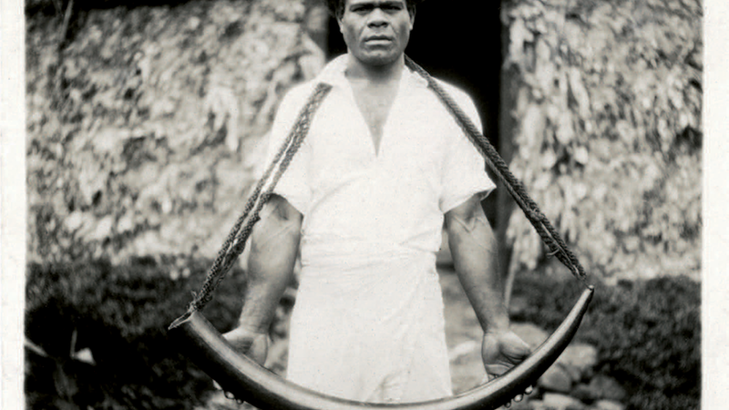 Sepia photograph of a man in white clothing holding an elephant tusk with a long cord.