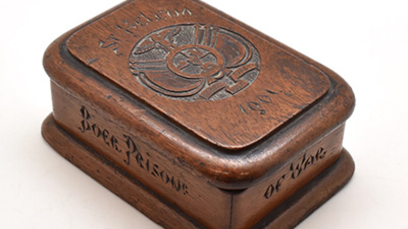 Rectangular wooden box with hinged lid. The top of the lid is inscribed with "St.Helena 1901" and depicts a crest including a bird, lion, and man with rifle; sides of box inscribed with "Made by Boer prisoner of war".