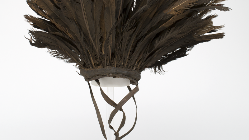 hat with long brown feathers mounted on a leather and cloth cap. Two chin straps attached to the edges.