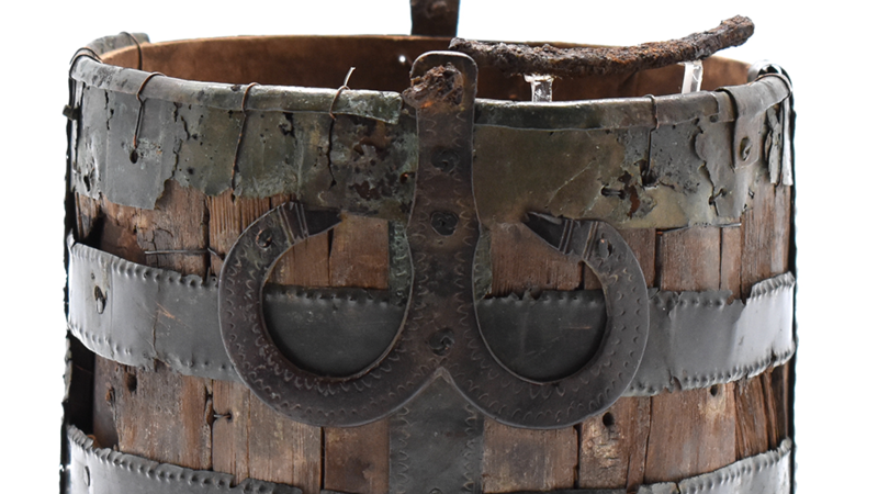 Cropped or close up image of the bucket, showing the weathered wood and hammered details of the metal bracing, with a double horseshoe shaped loop figure at the centre front.