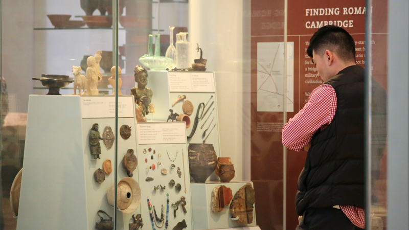A visitor stands in the local archaeology gallery looking at a case containing Roman finds, including metal figurines, pots, pieces of wall painting and jewellery.
