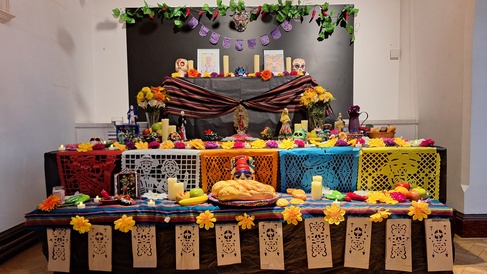 A 3-tier Day of the Dead altar draped in black fabric and brightly decorated with flowers, candles, food and paper cut outs.