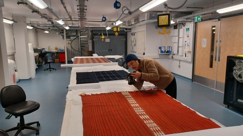 A workroom with three textiles laid out on a long table. A researcher with a camera is photographing a large red textile in the foreground with a red and cream pattern running along the centre.