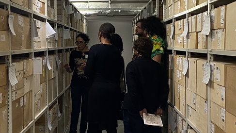 The advisory group stand between rows of shelves containing wooden boxes. The World Archaeology Curator talks to them about boxes containing material from the Caribbean.