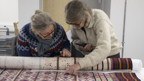 Two visitors to the museum's off-site store examine a partially unrolled textile. Both visitors point out different details in the making and design of the textile they're looking at.