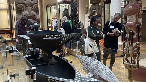 The advisory group explore the anthropology collections on display within one of the museum galleries. In the background rises a totem pole whilst in the foreground are large carved containers and a model of a shark.