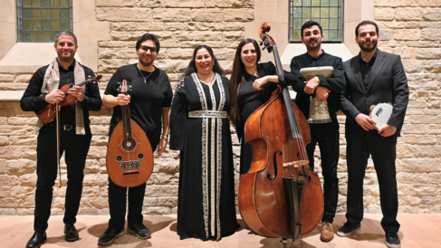 A group of six musicians stand in a line. Two hold percussion instruments whilst three others hold string instruments.