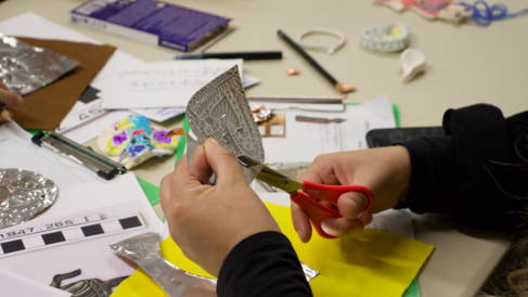A child, using a small pair of scissors, cuts around the embossed heart shape they've created. The table in front of them is covered in craft supplies and photos of objects within the collections.