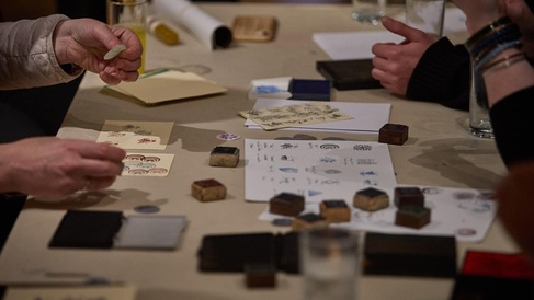 Three pairs of hands take part in a printing activity at a table within the museum galleries.