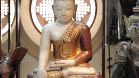 A seated statue of the Buddha on open display in the museum galleries. The Buddha sits crossed legged with a serene expression, one hand in his lap, the other touching the ground.