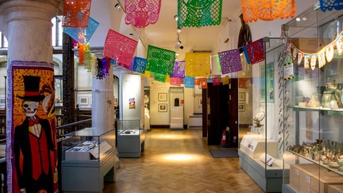 A long thin museum gallery decorated to celebrate Día de Muertos (Day of the Dead). There is brightly coloured bunting strung across the gallery and a depiction of a skeleton dressed in a three-piece suit and top hat attached to a column on the left.