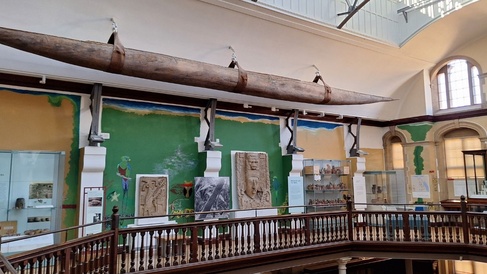 A museum gallery with a rail running around the centre of the room which is open to the gallery below. High on the wall hangs a long canoe and the gallery walls are decorated with mural featuring graffiti images relating to the collections on display.