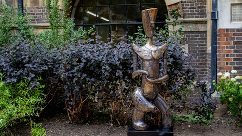 Photograph of the sculpture, a bronze carved form covered in symbols and patterns, on top of a black plinth in the MAA courtyard. The area is surrounded by green plants and the museum entrance is visible on the right hand side.