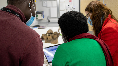 Rose Mwanja Nkaale, Nelson Abiti and MAA's Rachel Hand look at a computer screen showing the MAA database. A Ugandan object is visible on the table in the background; they are in the Centre of Material Culture, MAA's off-site storage facility.