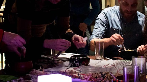 A group of people gathered around a craft table, painting gourds and other activities, at MAA. The lighting is dark and moody and there are drinks glasses on the table.