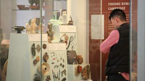 A visitor stands in the local archaeology gallery looking at a case containing Roman finds, including metal figurines, pots, pieces of wall painting and jewellery.