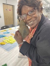 A smiling member of the advisory group holds a carved piece of stone up to her face. On the table behind her is a map covered in post-it notes.
