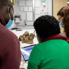 Rose Mwanja Nkaale, Nelson Abiti and MAA's Rachel Hand look at a computer screen showing the MAA database. A Ugandan object is visible on the table in the background; they are in the Centre of Material Culture, MAA's off-site storage facility.
