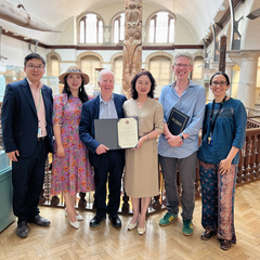 Six people, three women and three men, standing in the Museum of Archaeology and Anthropology, two of whom display a signed memorandum. They are smiling at the camera.