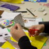 A child, using a small pair of scissors, cuts around the embossed heart shape they've created. The table in front of them is covered in craft supplies and photos of objects within the collections.