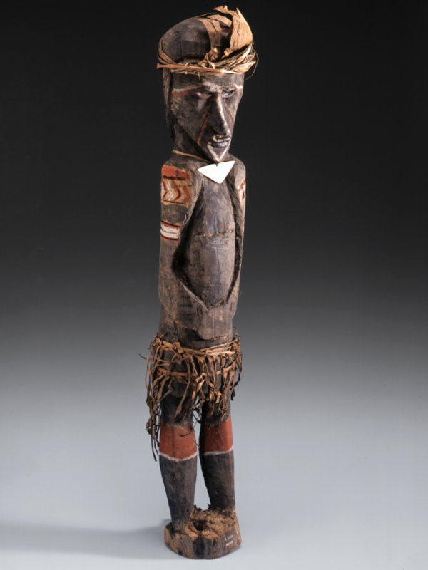 A carved wooden figure wearing a skirt of shredded leaf, a triangular chest ornament of pearl-shell, and palm-leaf head-dress.