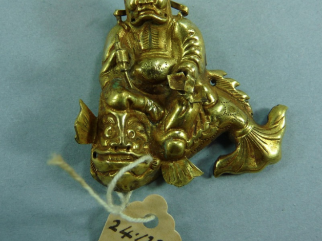 A copper gilt headpiece in the form of a frowning man seated on top of a fish. He has short horns and holds a writing brush in his right hand and a scholar's hat (or peck measure) in his left.