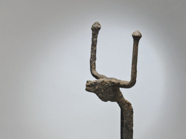 Detail of a bronze horned animal head at the top of a firedog, against a grey studio background.