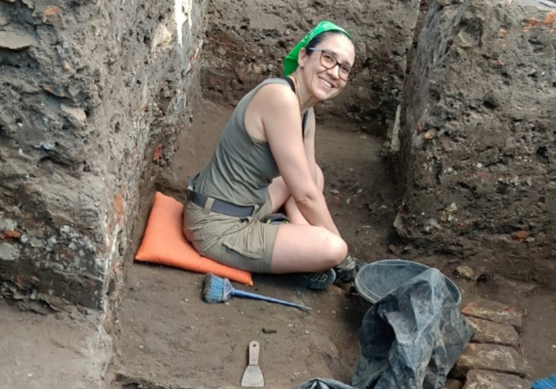 The museum's World Archaeology Curator sits in the bottom of a trench and smiles up at the camera.