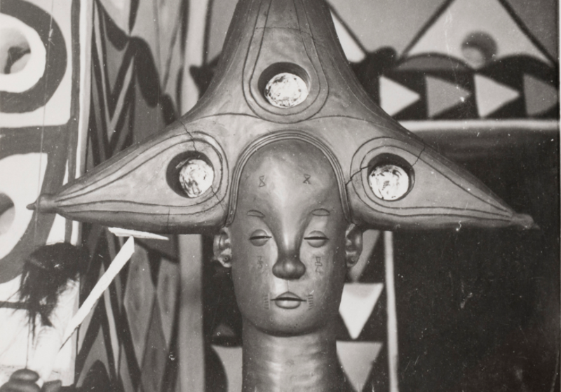 A close up of the clay head of a female deity. She has a three-horned coiffure, closed eyes and mouth and a broad nose. 
