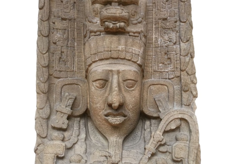 The face of a king which looks like it has been carved from stone. He wears large ear ornaments and a large headdress, which has two plumes of feathers running down the sides.