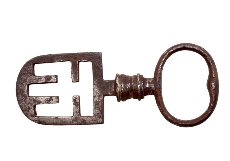 A metal key with an oval bow at one end, a short baluster-like stem, and a symmetrical openwork spade-shaped bit with four internal wards at the other end.