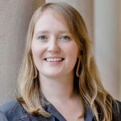 A curator with long blond hair smiles. She wears large dangling earrings and has a small nose piercing.