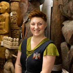 Staff photograph of Katrina Dring, smiling at the camera, wearing a green top and black denim overalls, with a colourful beaded badge. She stands in front of a collection of statues, nestled in the corner of the MAA Stores.