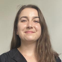 A woman with long brown hair and wearing a black blouse smiles at the camera. Behind her is a pale coloured wall and the ends of shelving to the left of the image.