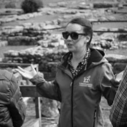 A black and white photo of a woman with dark hair and sunglasses in front of excavated ruins.