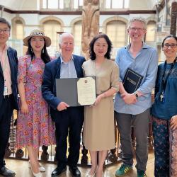Six people, three women and three men, standing in the Museum of Archaeology and Anthropology, two of whom display a signed memorandum. They are smiling at the camera.
