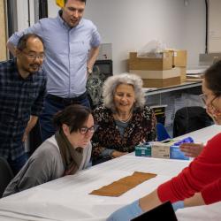 Six people, both sitting and standing, peer over a barkcloth with interest and intrigue, that is being presented to them by Emily and Jane who wear red MAA jumpers. The barkcloth is medium sized, and laid out on conservation paper on a workshop table.