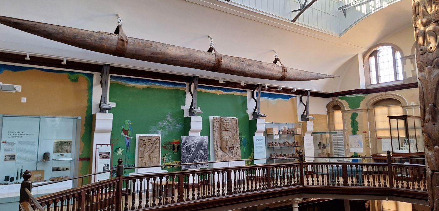 A museum gallery with a rail running around the centre of the room which is open to the gallery below. High on the wall hangs a long canoe and the gallery walls are decorated with mural featuring graffiti images relating to the collections on display.