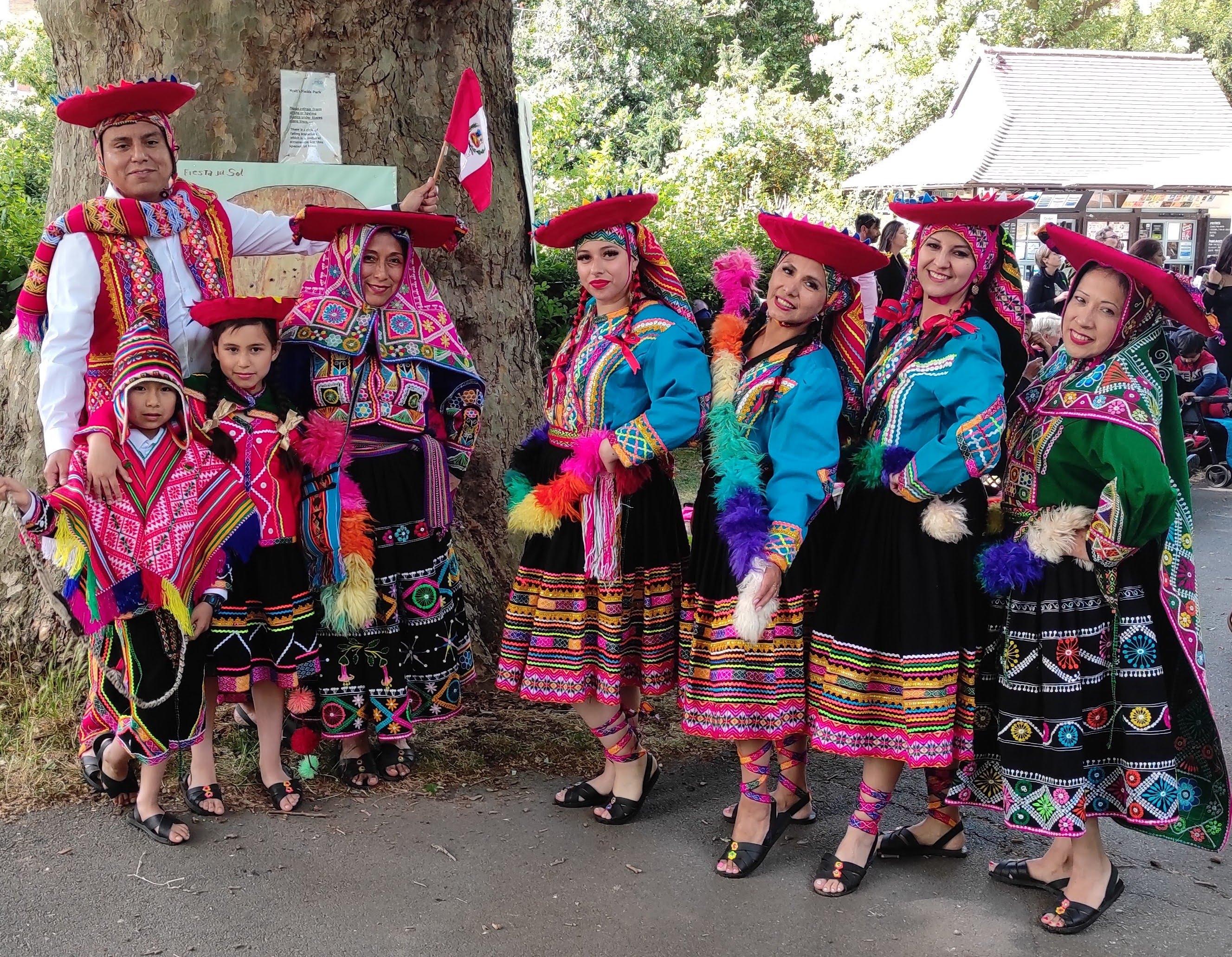 A group of six adults and two children stand outside, under a tree. They are dressed in brightly coloured Peruvian clothes and most of them are wearing red hats.