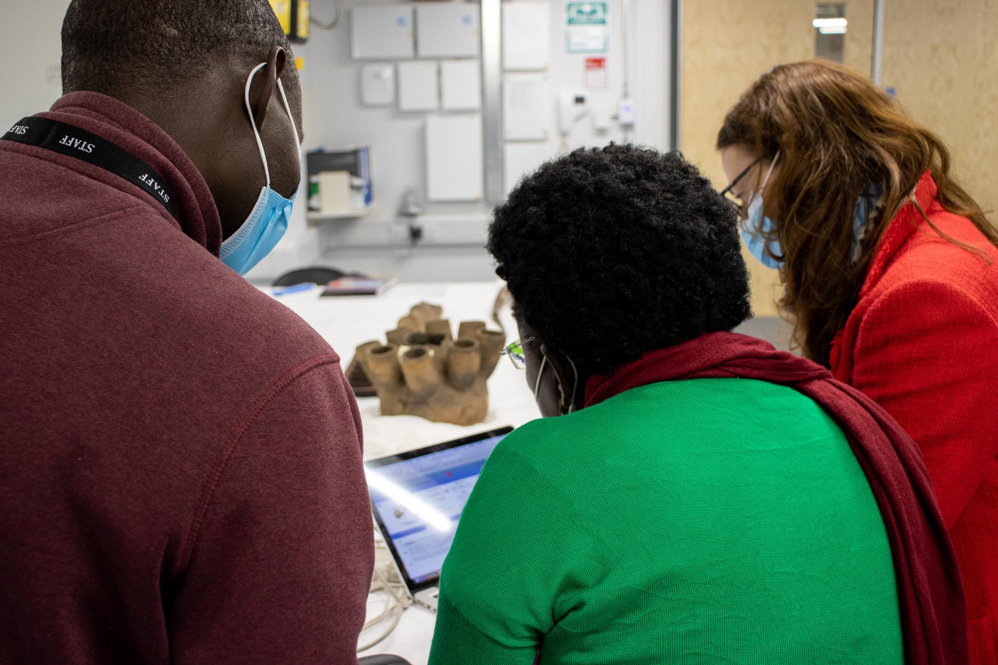 Rose Mwanja Nkaale, Nelson Abiti and MAA's Rachel Hand look at a computer screen showing the MAA database. A Ugandan object is visible on the table in the background; they are in the Centre of Material Culture, MAA's off-site storage facility.