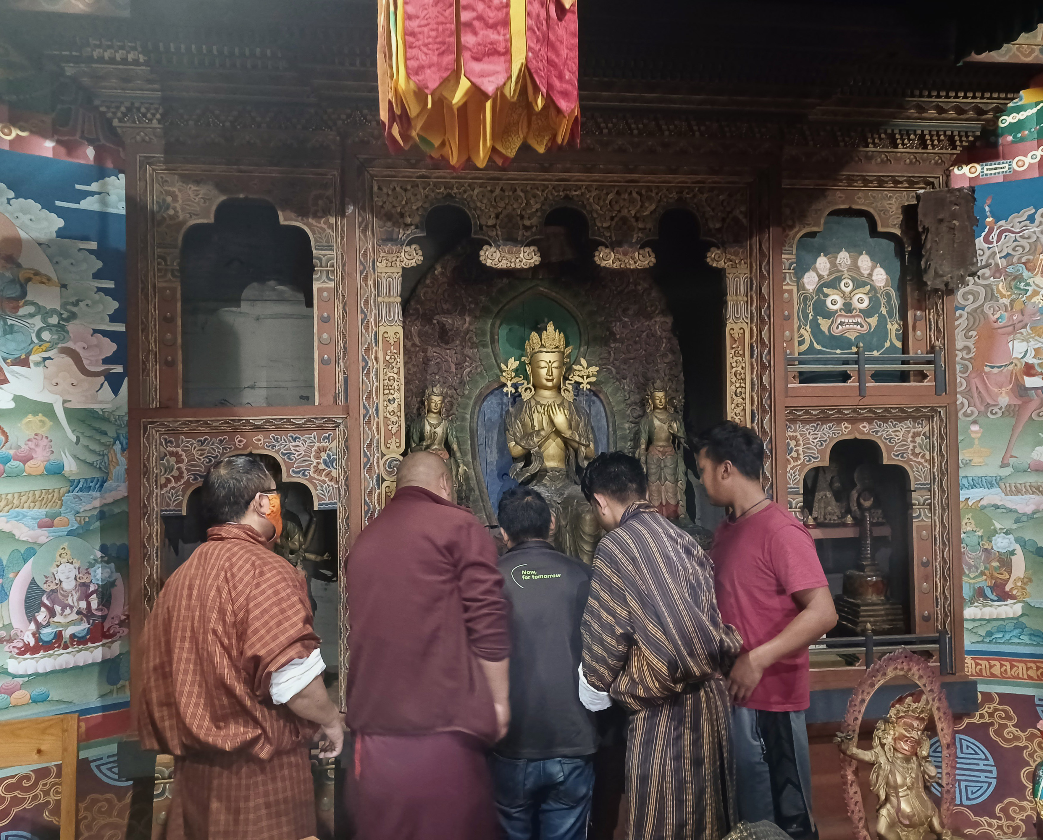A group of five men with their backs to the camera grouped in front of a shrine. This contains three gold coloured statues, the central one is the larger than the others. The shrine is highly decorated and the walls are painted with religious scenes.