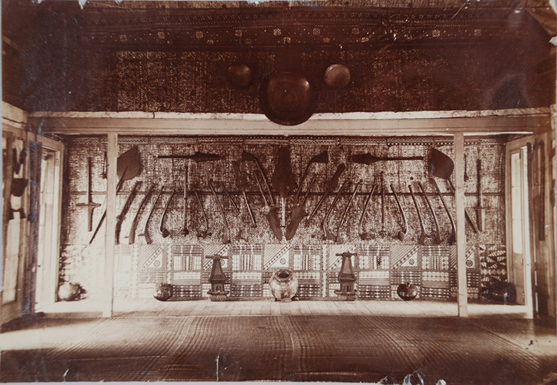 Sepia photograph of a large room, with objects hung along the back wall and from the ceiling. The room is empty aside from them.
