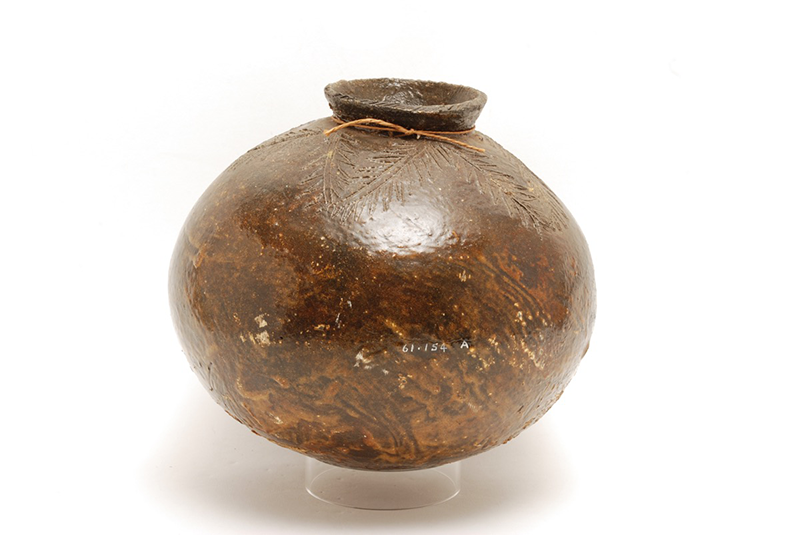 Rounded water pot in a mid-brown, decorated with some leaf pattern at the top but otherwise undecorated. Simple opening at the top with a small cord/label tag attached.