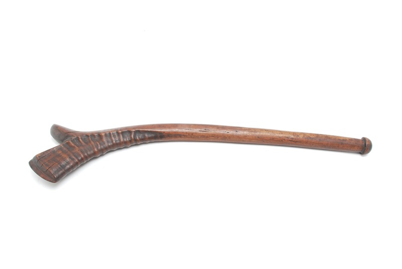 A mid-dark wooden club. The head is curved and the sides are carved in waves of undulated ridges.
