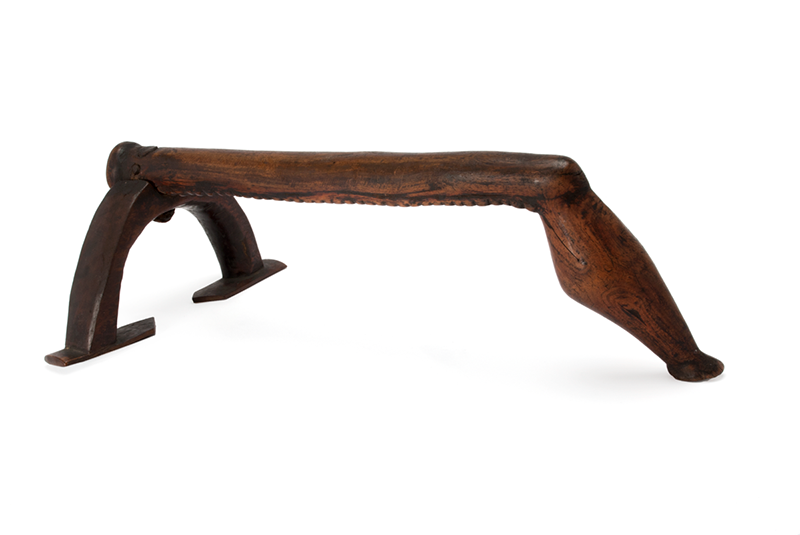 Dark wood headrest with a piece of wood curving down to form one of the legs and carved in the shape of a leg.