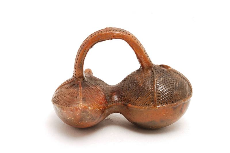 Vessel made of two connecting circular pods, the right larger than the left, connected centrally and with an arched handle. The top half is decorated with incisions and dots. Pouring spout from the larger end. Glazed, red-brown in colour.